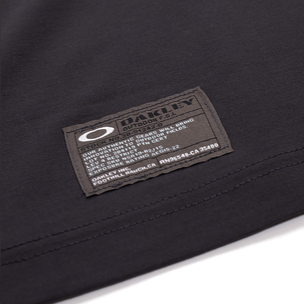 Macro studio view of the hem branding on the Oakley FGL Union SS Tee 6.7 in Phantom, showcasing the stitched dark grey technical spec label with the white Oakley Ellipse logo, "OAKLEY OUTDOOR F.G.L." insignia, and field-issue product parameters.
