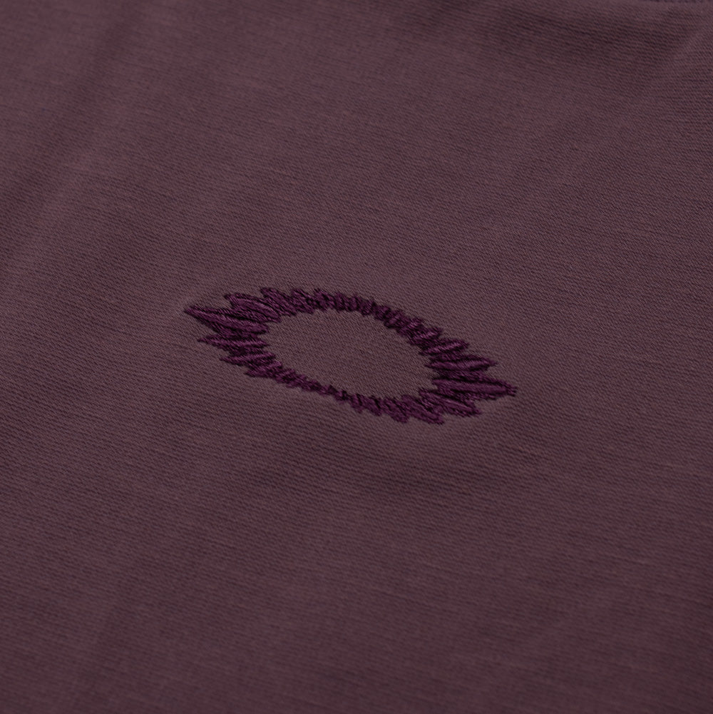 Macro studio close-up of the chest embroidery on the Oakley FGL Union SS Tee, highlighting the tonal deep plum thread forming a jagged, soundwave-style embroidered outline of the Oakley Ellipse emblem on smooth technical jersey knit fabric.
