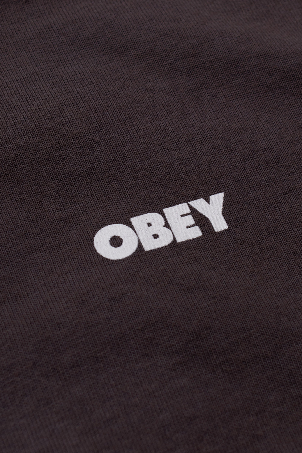 Macro detail view of the clean white Obey bold logo text printed across the textured pigment-dyed dusty black cotton jersey fabric on the front chest.