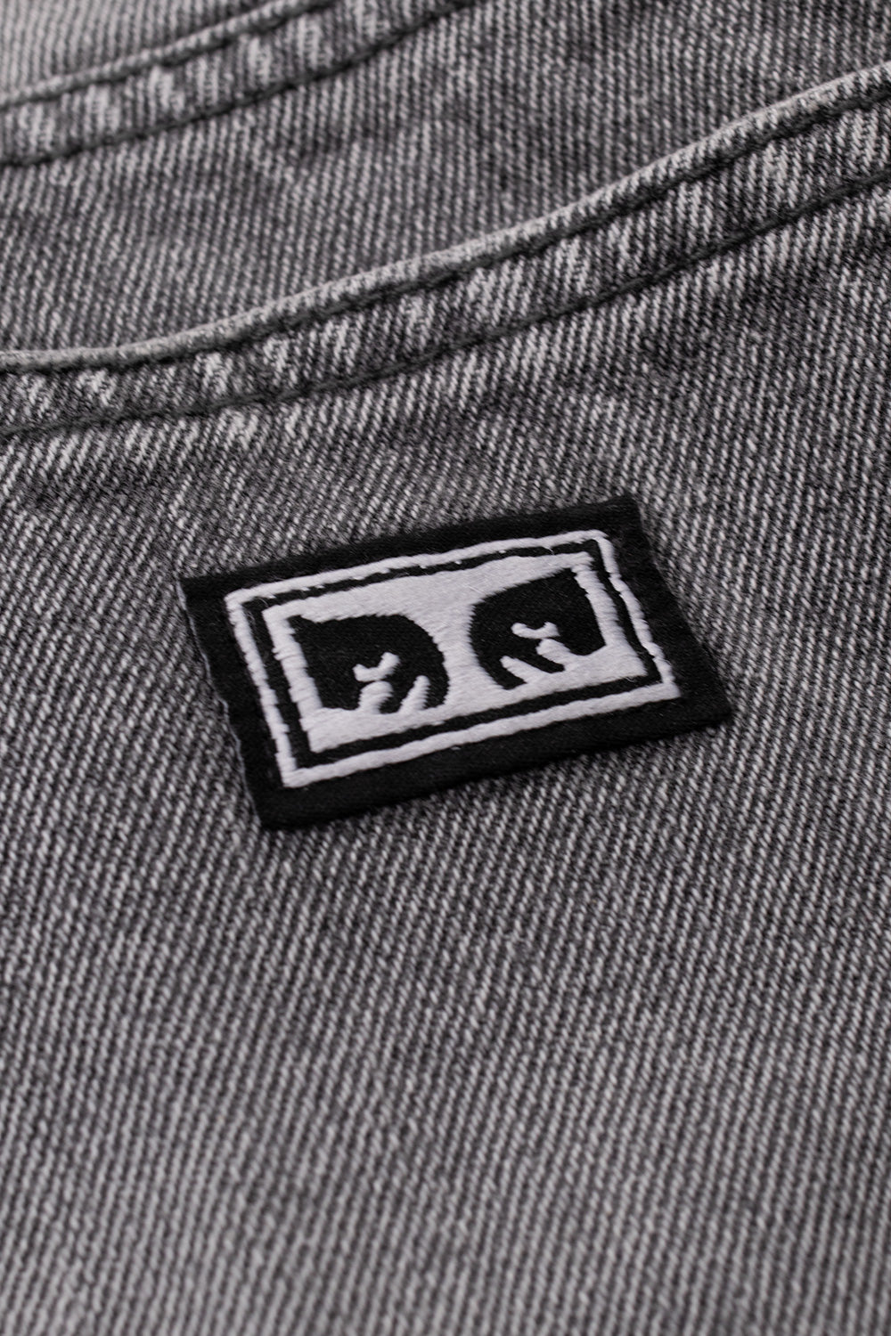 Close-up detail shot of the back right pocket on the Archive Grey Obey Boxcar jeans, highlighting the twill texture of the washed 13.5oz cotton denim and the woven black and white Obey André the Giant Eyes brand patch with clean border stitching.