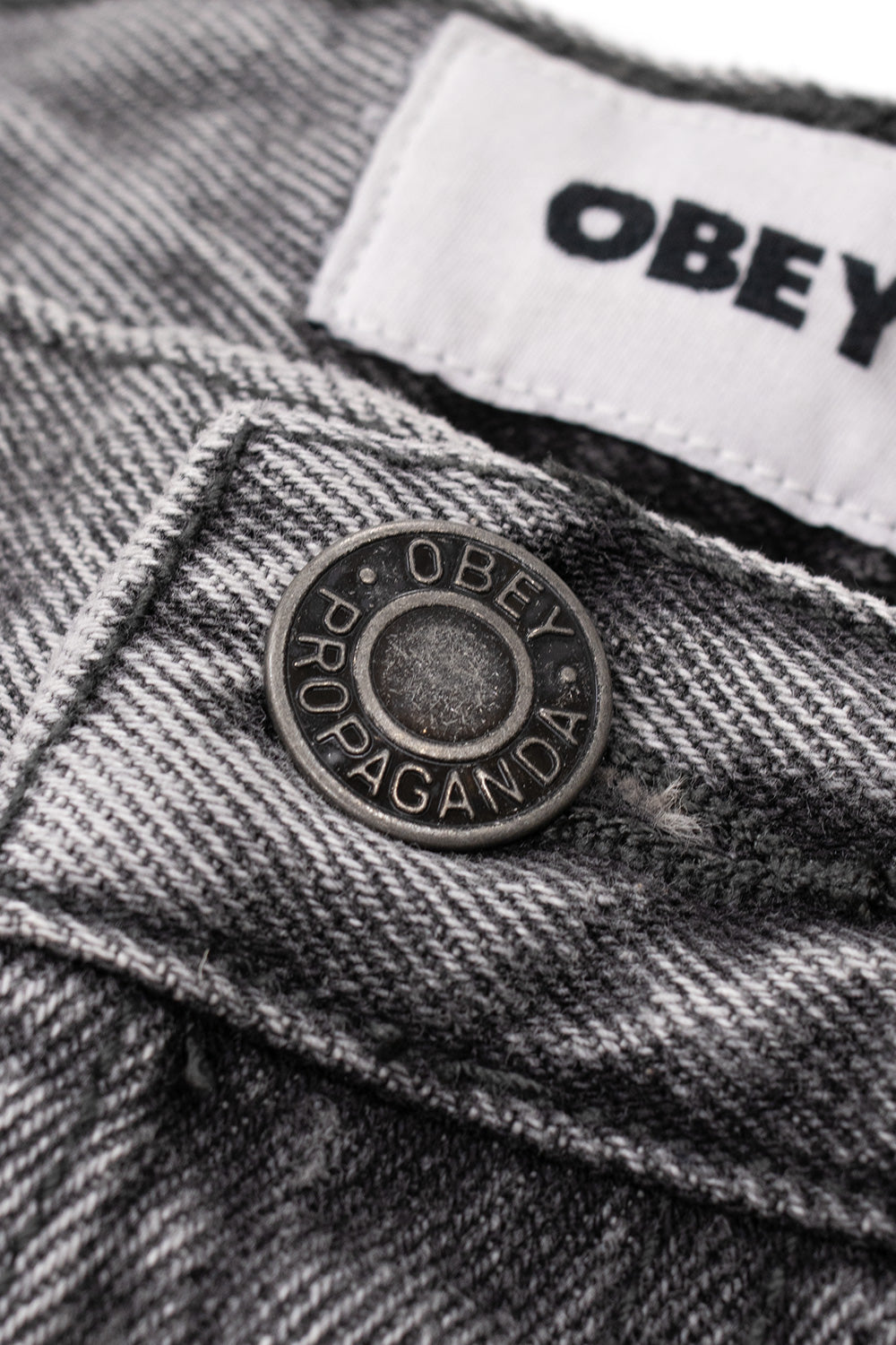 Macro detail shot of the waistband closure on the Obey Boxcar denim, displaying the embossed antique metal button inscribed with Obey Propaganda typography set against the Archive Grey twill denim and white woven inner Obey brand label.