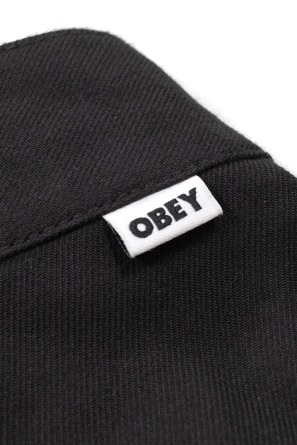 Macro detail shot of the back waist on the black Obey Dean Pleated Pant, showing the woven diagonal texture of the durable black poly-rayon twill fabric and the small white rectangular Obey flag label with bold black typography.