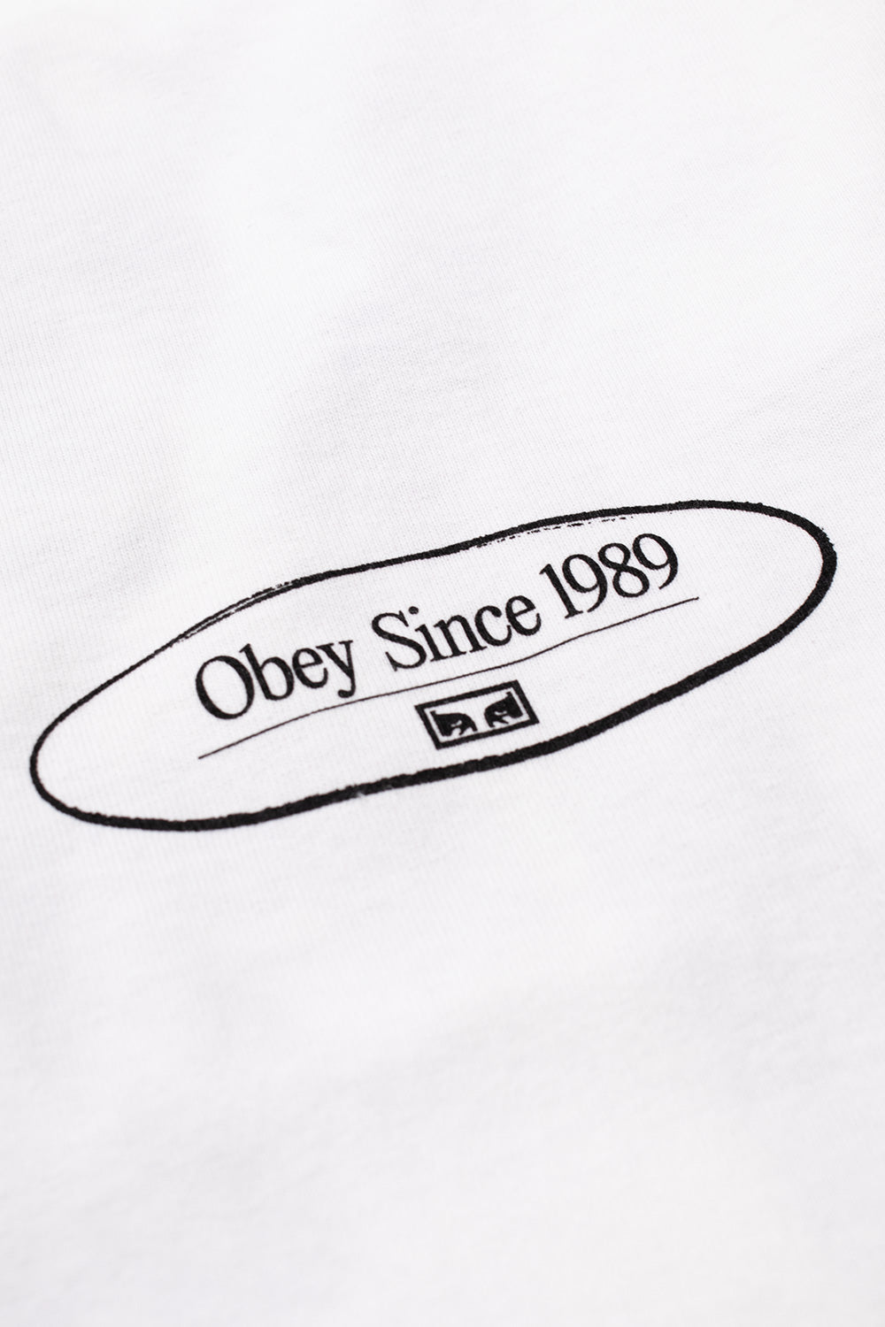 Macro detail view of the black oval Obey Since 1989 chest logo print featuring the iconic miniature Shepard Fairey Eyes emblem on the fine white cotton jersey fabric.