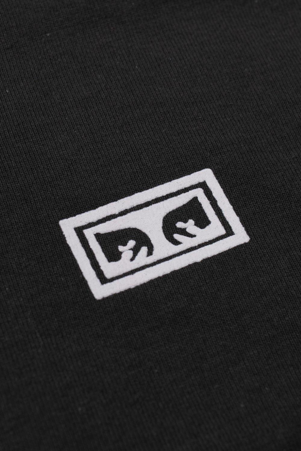 Macro detail view of the miniature white Obey Eyes box logo printed on the textured vintage black cotton jersey fabric on the front chest.