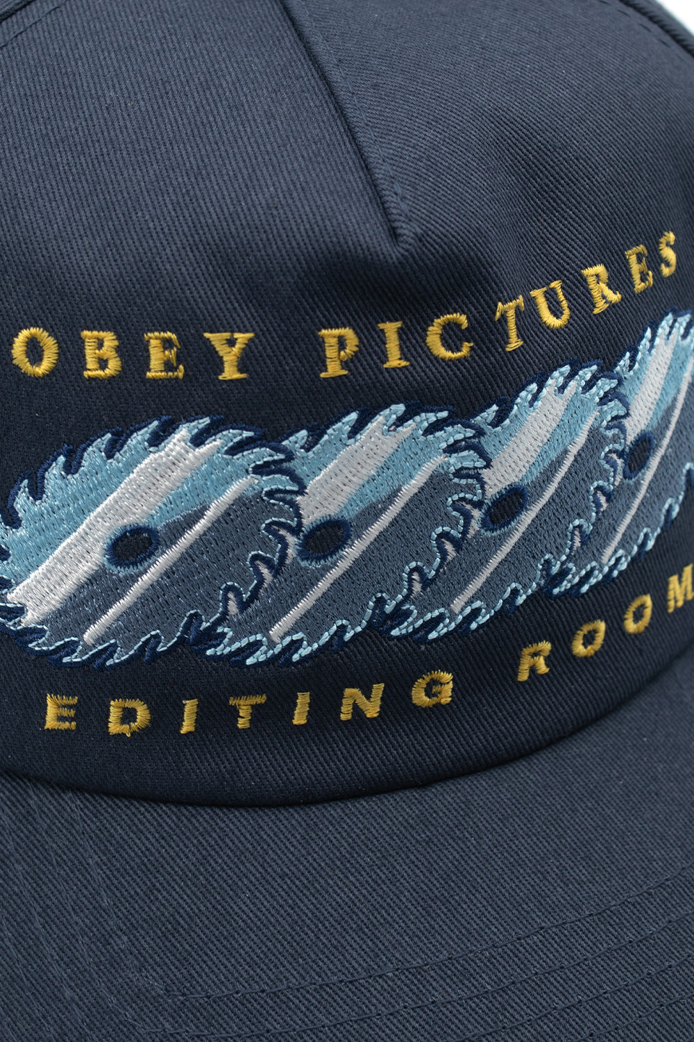 Macro close-up shot of the intricate embroidery on the navy Obey Pictures cap, detailing the golden-yellow stitched Obey Pictures Editing Room lettering surrounding dynamic overlapping circular saw blade graphics in white, light blue, and navy thread.