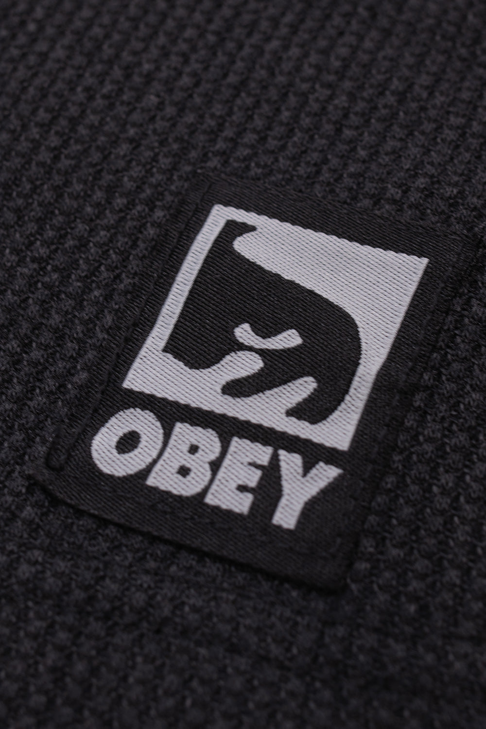 Macro close-up shot of the lower left hem on the Dusty Black Limo Obey Post Thermal, displaying the three-dimensional square grid texture of the coarse waffle knit fabric and the sewn-on black and grey woven Obey patch featuring the iconic stylized André the Giant graphic and bold serif Obey lettering.