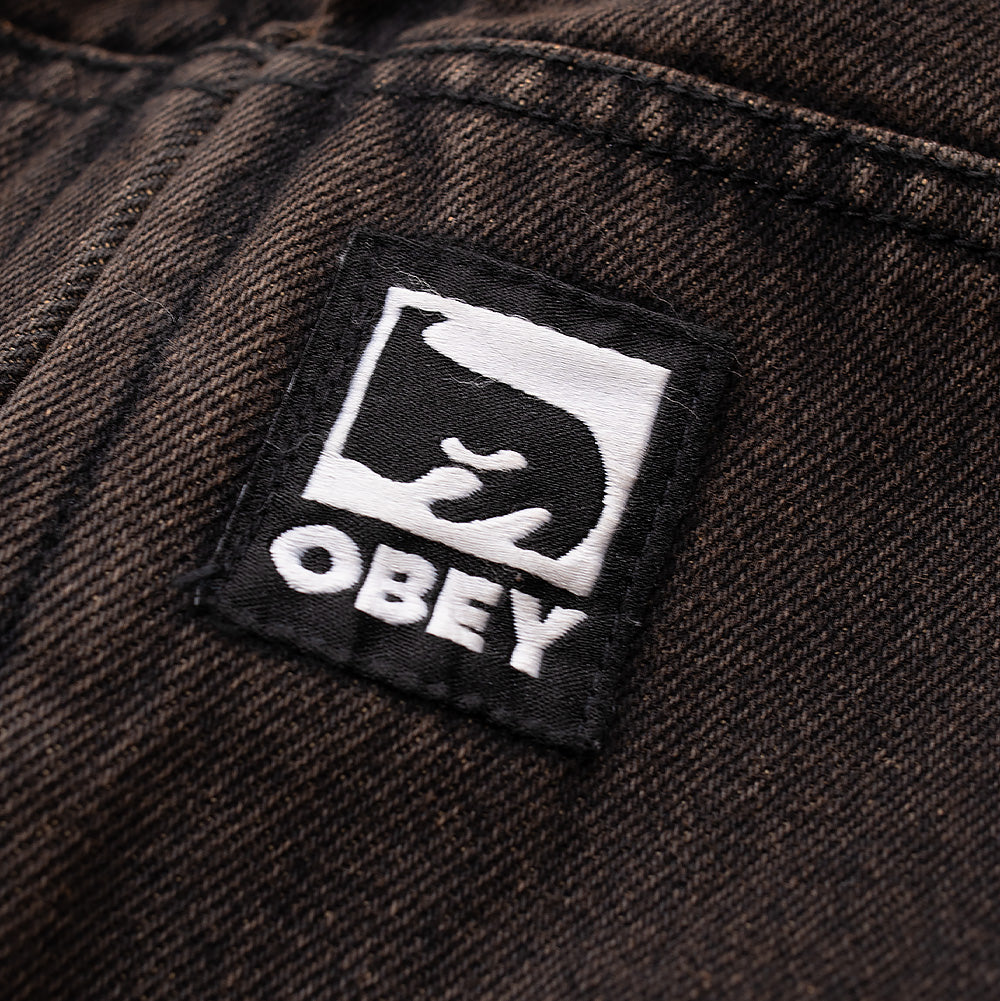 Detailed close-up shot of the woven square brand patch on the Obey Studded Zip Up Jacket, showcasing the iconic black and white Obey Icon Eyes graphic and bold "OBEY" block lettering sewn onto the diagonal twill of the faded rust-washed pocket seam.