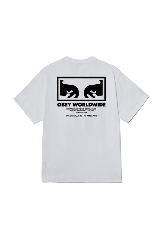 Back view of the white Obey The Medium Is The Message T-Shirt, crafted from 100% cotton jersey, featuring a bold black screen-printed Shepard Fairey Giant Eyes box graphic, Obey Worldwide lettering, international city names, and The Medium Is The Message slogan.