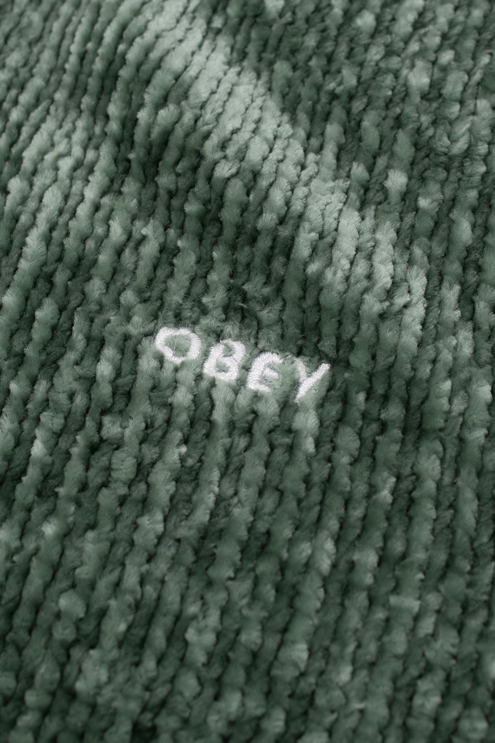 Close-up view of the crisp white embroidered Obey script logo on the textured Dark Forest green ribbed chenille fabric on the left chest panel of the Varen Zip Up Sweater.