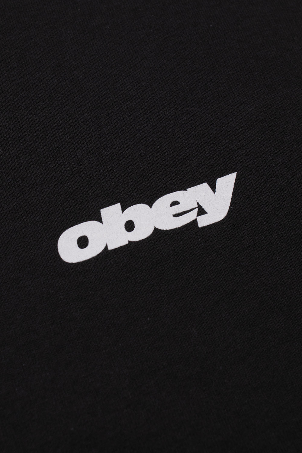 Macro detail view of the solid white Obey italic bold text logo printed on the fine black 100% cotton jersey fabric on the front chest.