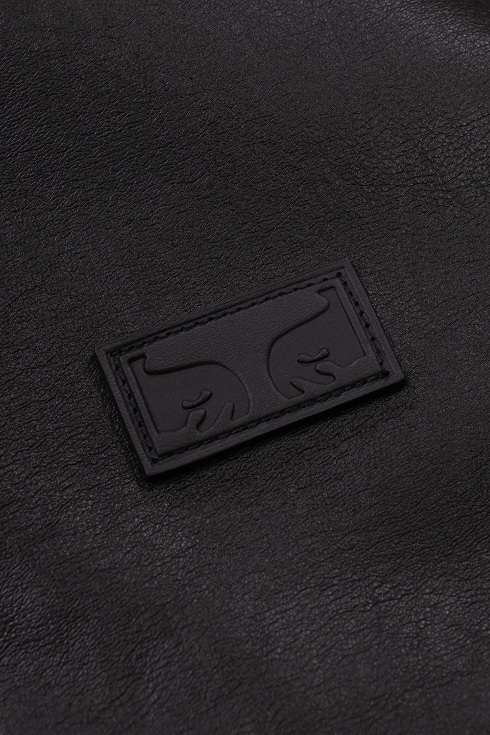 Macro close-up shot of the black faux leather chest patch on the Obey Workman PU Jacket, featuring stitched perimeter borders and debossed Shepard Fairey Giant Eyes logo branding.