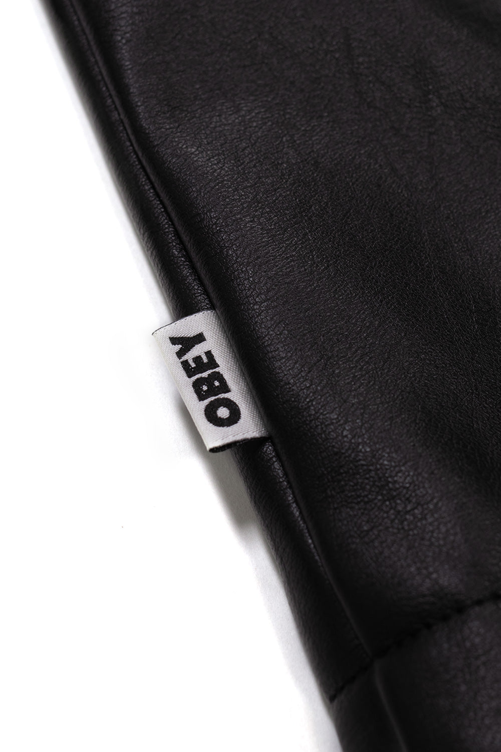 Close-up shot of the white woven Obey flag label stitched into the side seam of the black Workman PU leather jacket.