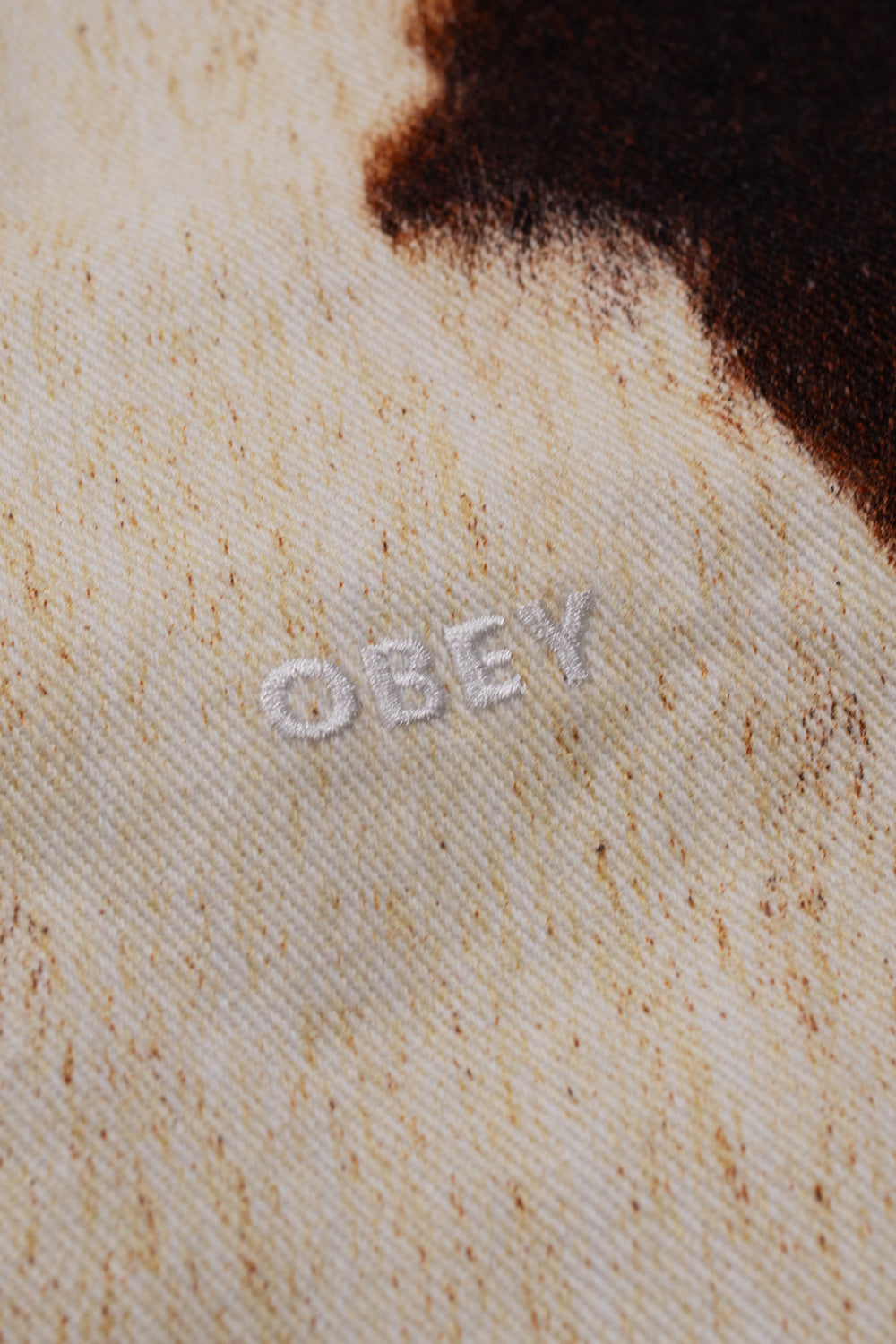 Detailed view of the fine white embroidered Obey logo stitched into the heavyweight textured twill cotton fabric with realistic brown cow-fur print on the jacket chest.