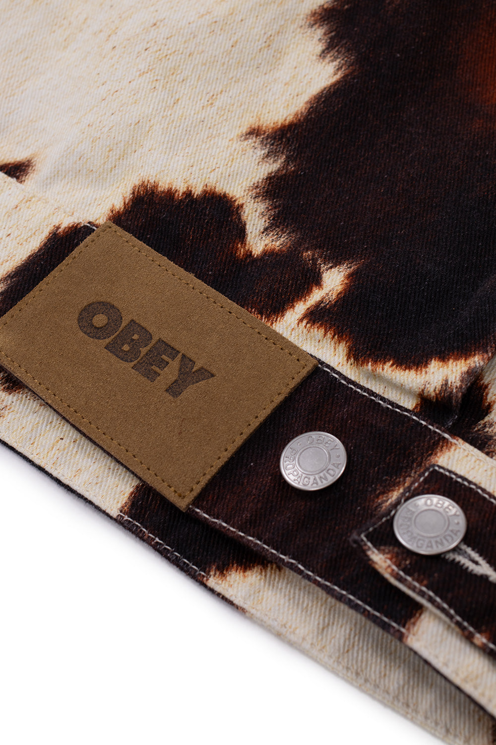 Macro detail shot of the lower back hem on the Obey Workshop Jacket, showing the embossed brown suede leather Obey logo patch alongside branded silver metal Obey Propaganda shank buttons on the adjustable waist tab.