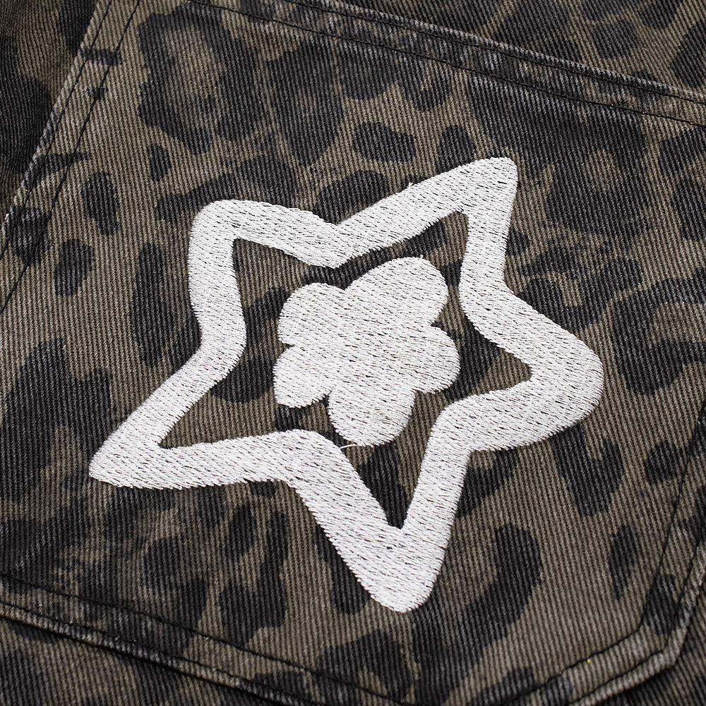 Macro close-up shot of the rear right pocket embroidery on the Starteam East Village Leopard Star Jeans, showcasing the dense, raised white chain-stitch embroidery outlining a five-pointed star surrounding a central blooming flower motif over the muted dark olive leopard denim weave.
