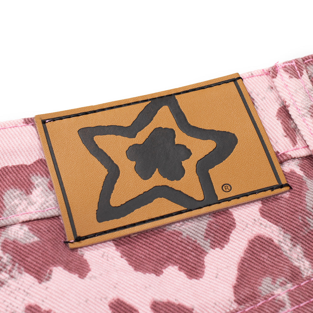 Macro studio shot of the rear waistband jacron patch on the Starteam East Village Leopard Star Jeans in Pink, displaying the smooth tan faux-leather label debossed with the thick black outline of the Starteam flower-star graphic and registered trademark symbol, firmly sewn onto the pink leopard denim waist.