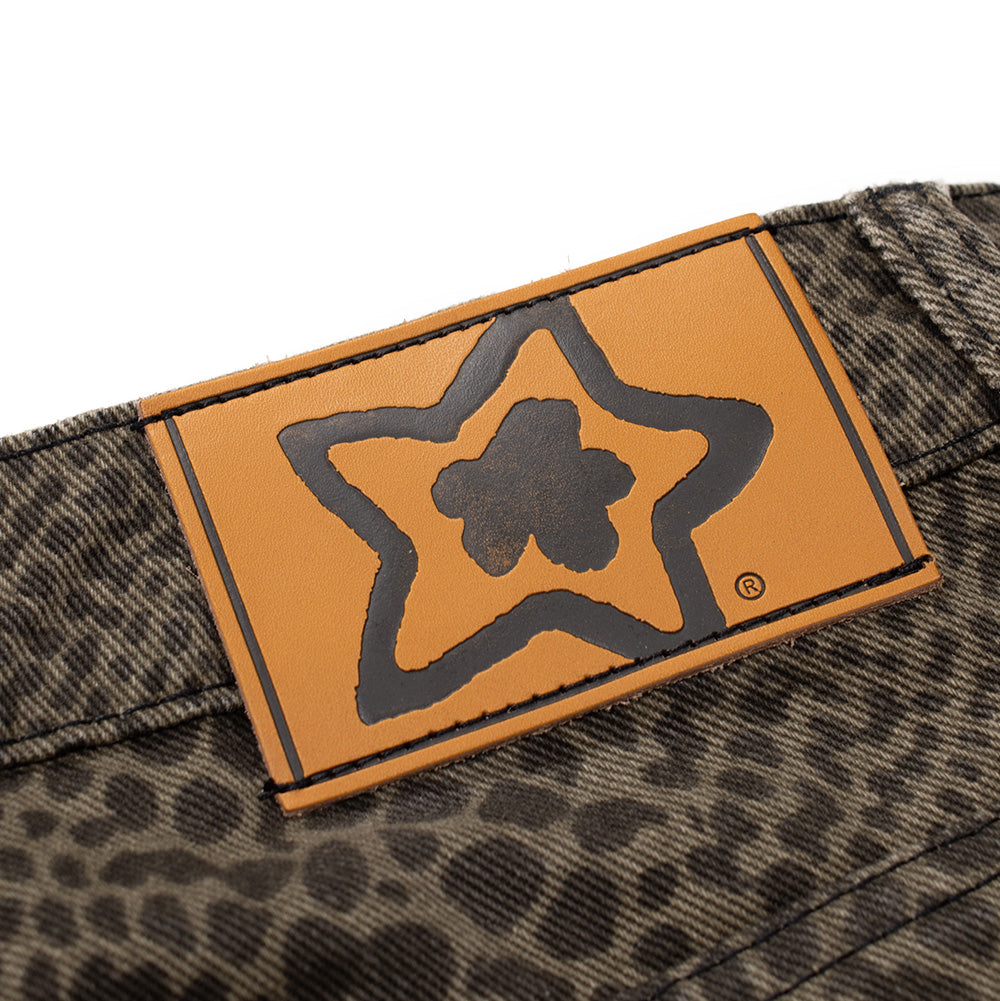 Macro studio shot of the rear waistband jacron patch on the Starteam East Village Snakeskin Star Jeans, displaying the light cognac brown faux-leather label debossed with the thick black outline of the Starteam flower-star emblem and registered trademark symbol, stitched onto the textured olive snakeskin denim.