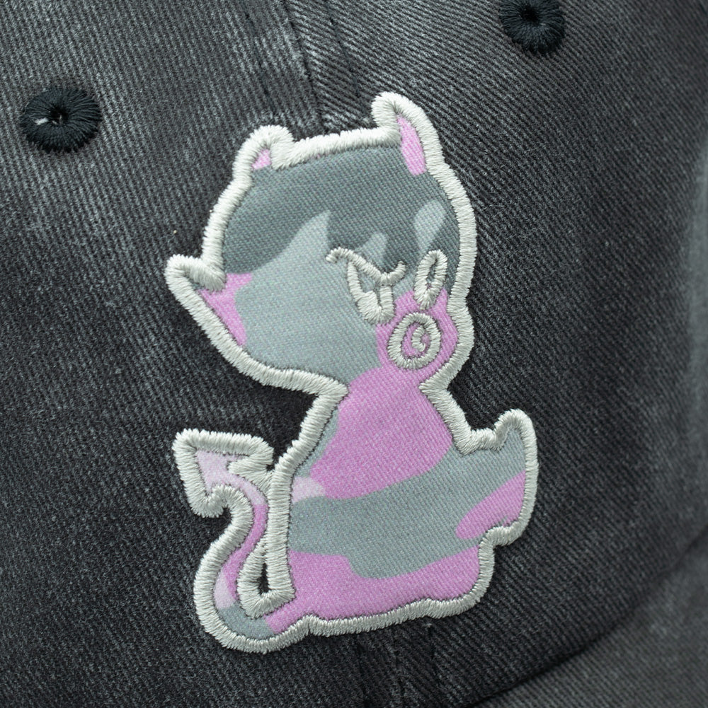 Detailed macro studio close-up of the front patch on the Token Bae Cap, showing the thick white satin-stitched perimeter and the printed pink, grey, and white camouflage fabric featuring an illustrated mischievous cartoon devil character with horns, tail, and pacifier.