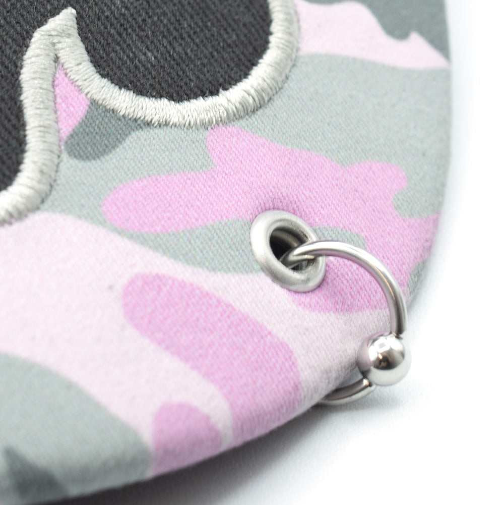 Extreme macro shot of the curved visor on the Token Bae Cap, highlighting the silver metal grommet eyelet inserted through the pink camouflage fabric holding a captive silver metallic ball-closure piercing hoop ring.