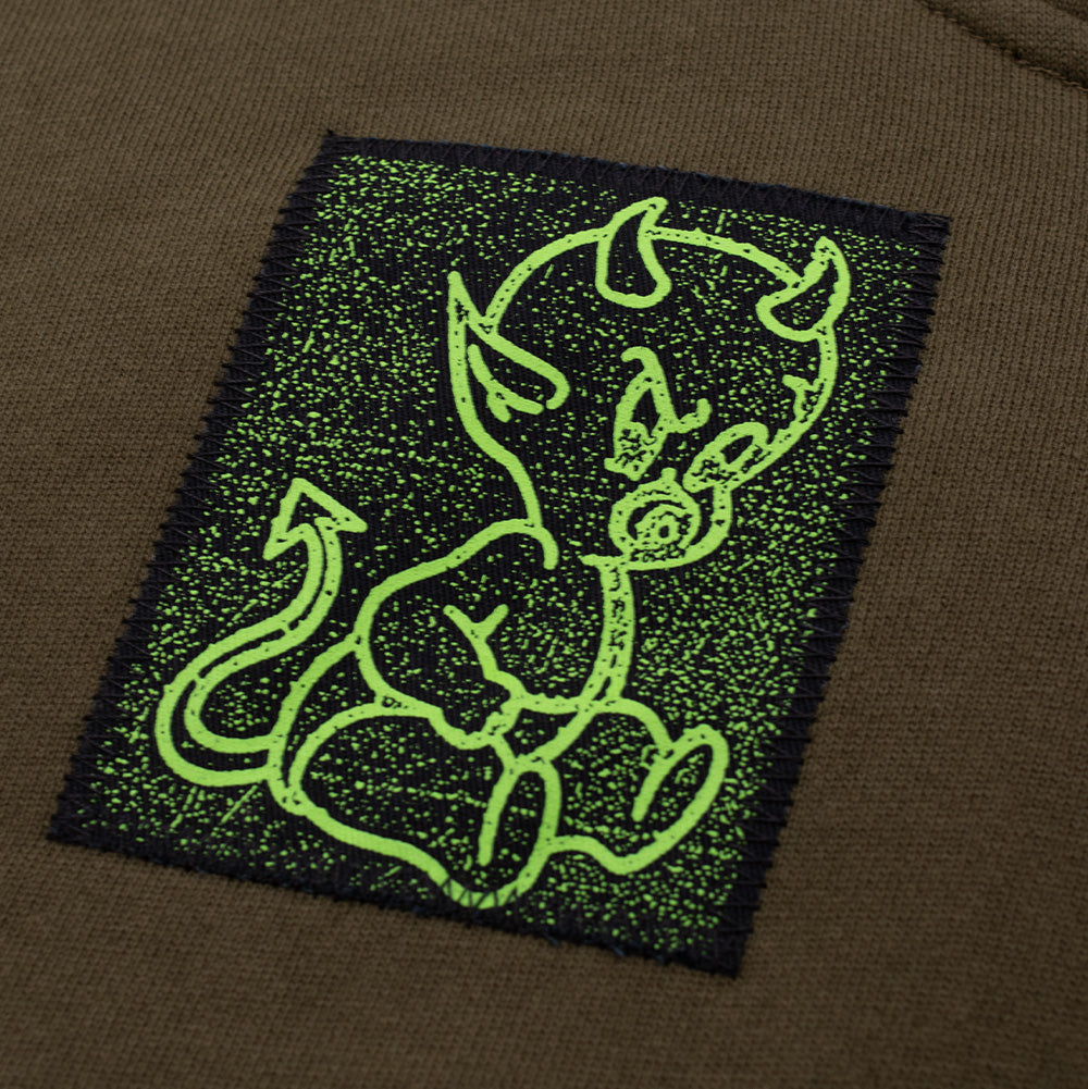 Macro studio view of the upper right chest patch on the Token Carly Zip Hoodie, showing the raw frayed canvas edges secured with black zigzag stitching and a lime neon green screen print of a mischievous baby devil cherub on a black background.