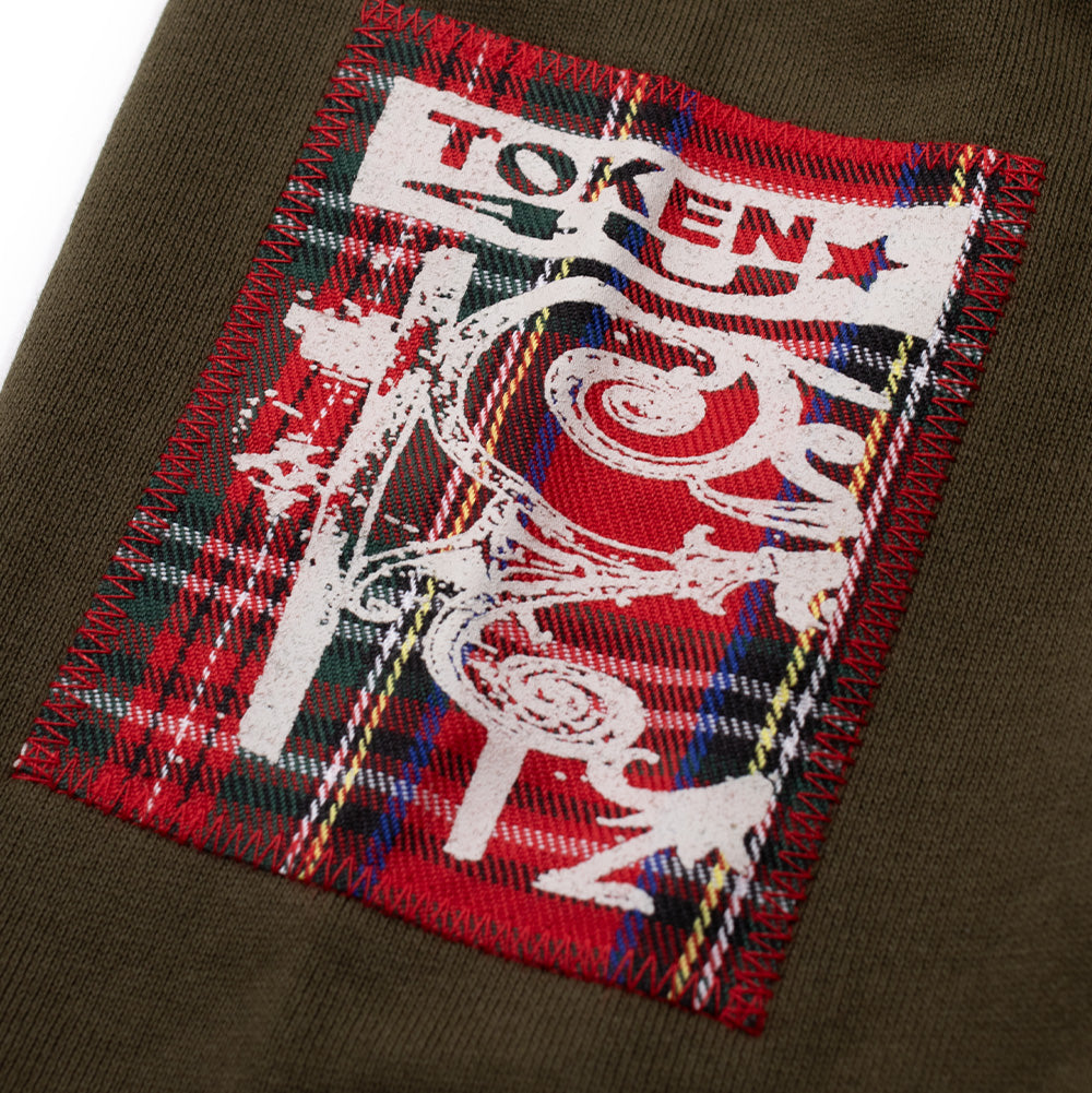 Macro close-up perspective of the right sleeve cuff patch on the Token Carly Zip Hoodie, highlighting a red tartan plaid fabric square cross-stitched in red thread, printed with an off-white street lamp illustration and the bold "TOKEN" logo with a red star.