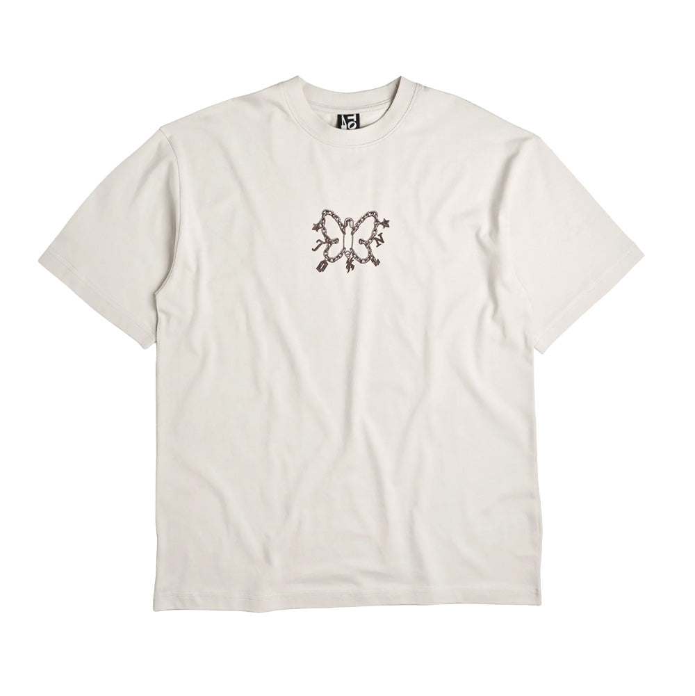 Front flat lay view of the off-white Token Expansive Skies T-Shirt, featuring a relaxed skate fit, ribbed crewneck collar, short sleeves, and a small dark brown center-chest graphic print shaped like a butterfly formed from link chains, charms, letter pendants, and a large safety pin body.