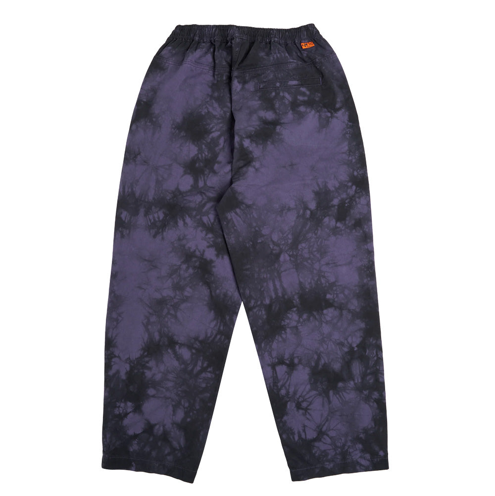 Back flat lay view of the Token Sundown Pants in purple black tie dye, showcasing the tapered roomy fit, seamless back yolk, rear welt pocket, elastic waistband, and a small bright orange woven Token brand logo label sewn on the upper right hip seam.