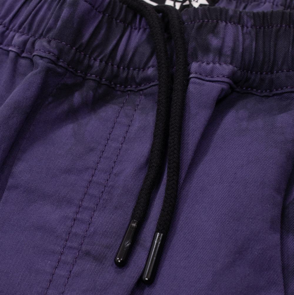 Macro studio close-up of the gathered elastic waistband on the Token Sundown Pants, highlighting the dark purple cotton twill fabric, tonal stitching, and black round drawstring cords with coated aglets emerging from the front.