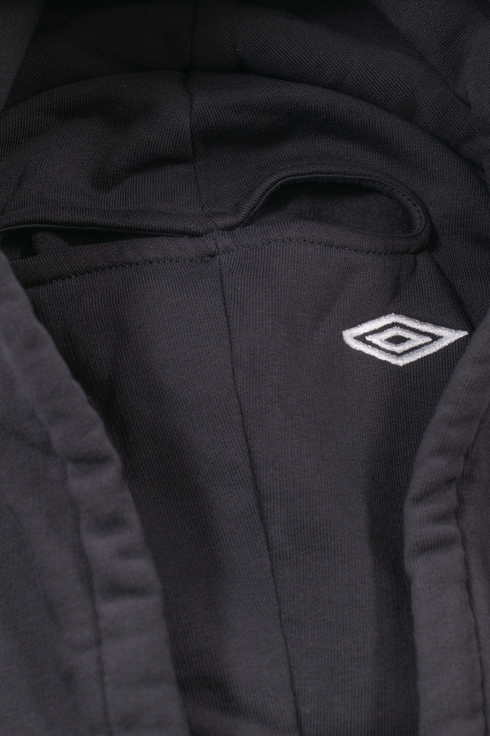 Macro close-up shot of the hood on the Umbro Authority Zipped Hoodie, displaying the integrated black cotton jersey face mask with a bound horizontal eye opening, accented by a white embroidered Umbro double diamond logo on the cheek area