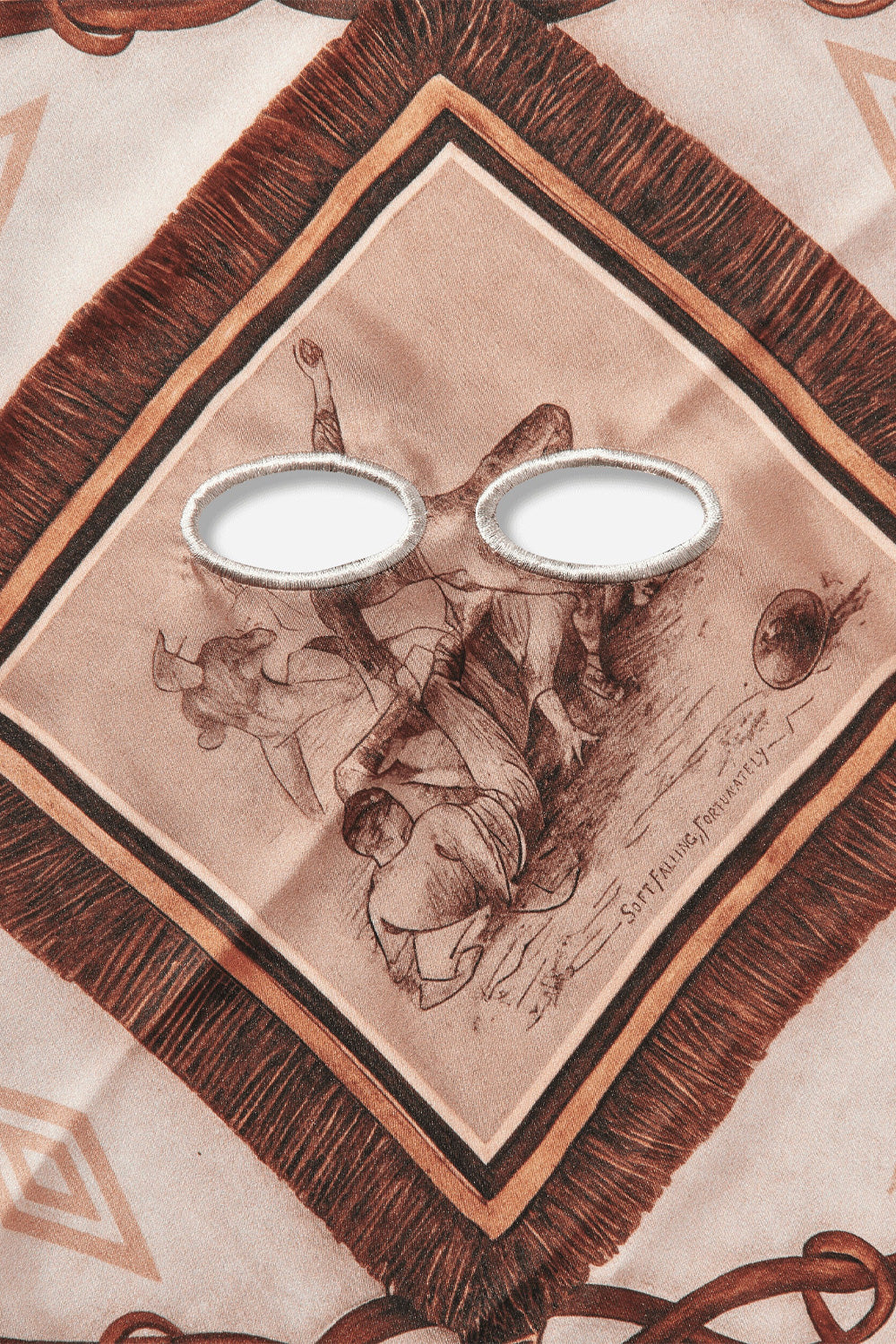 Detailed close-up shot of the central diamond frame on the Umbro Belt Masked Foulard, showcasing two oval cut-out eye openings finished with cream satin-stitched embroidery over a sepia illustration of football players sliding for a ball, captioned "SOFT FALLING, FORTUNATELY" and bordered by printed brown fringe tassels
