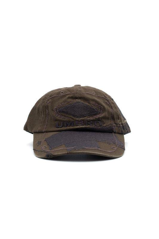 Front studio portrait of the Umbro Censored Cap in washed dark olive-brown cotton, displaying the curved visor and unstructured 6-panel crown embellished with tonal ornamental scroll embroidery, an embroidered black Umbro wordmark, and raw-edged distressed fabric patches stitched across the peak and front panels to censor the central logos