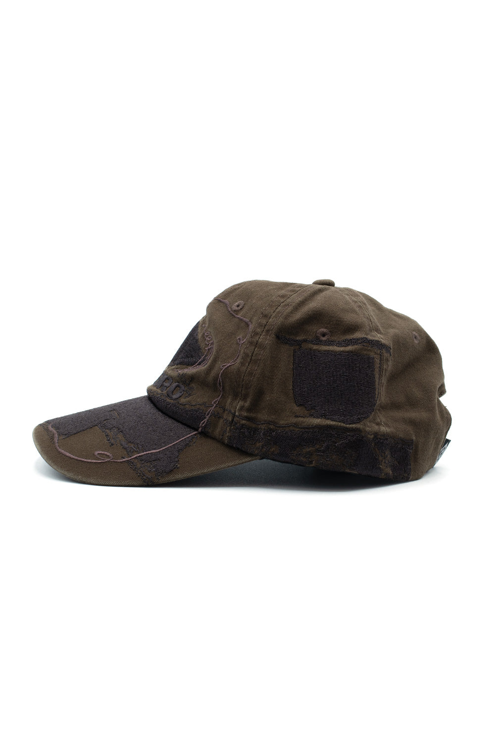 Left side profile view of the Umbro Censored Cap, highlighting the low-profile crown shape, curved brim, continuous decorative embroidery extending along the side panel, and raw-edge shield-shaped and rectangular distressed fabric patches sewn onto the cotton body