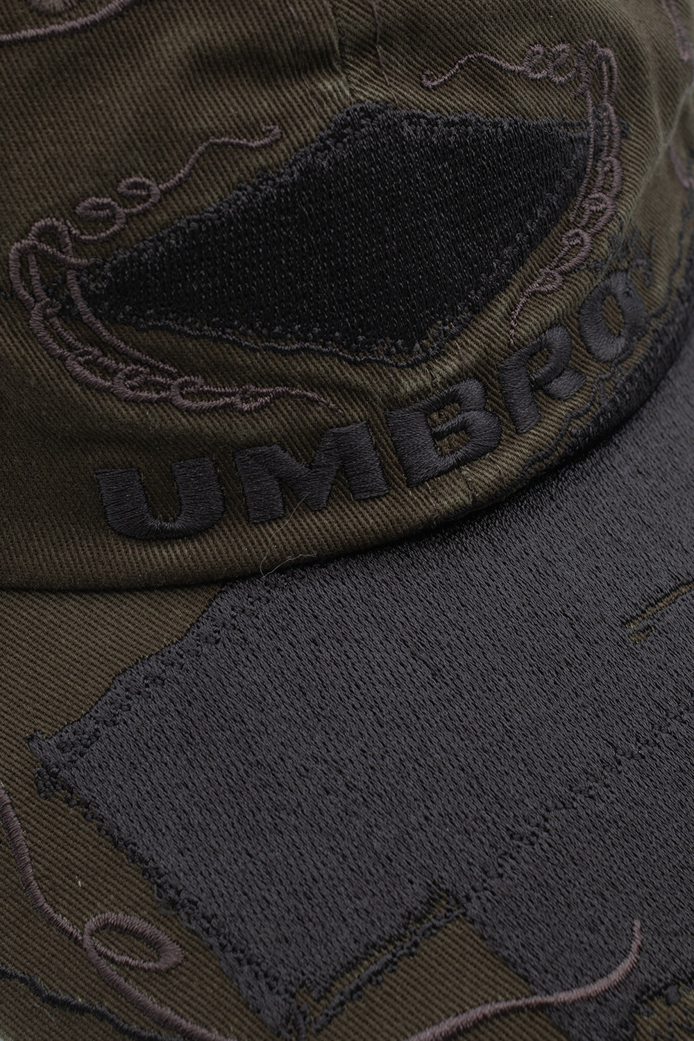 Macro close-up shot of the front panel and visor on the Umbro Censored Cap, detailing the dense black satin embroidery of the Umbro wordmark beneath an embroidered diamond patch, fine baroque threadwork scrolls, and heavy zig-zag edge stitching on the raw-cut fabric overlays