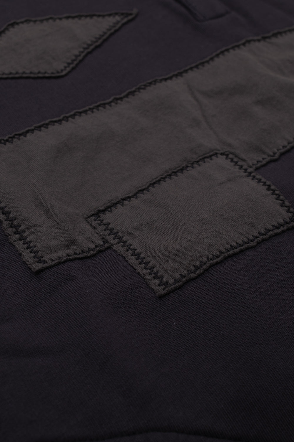 Macro studio close-up of the chest application on the Umbro Censored Pearls Hoodie, showing raw-edged black cotton jersey fabric patches sewn in diamond, shield, and horizontal bar shapes using thick contrast black zig-zag stitching over the washed fleece fabric