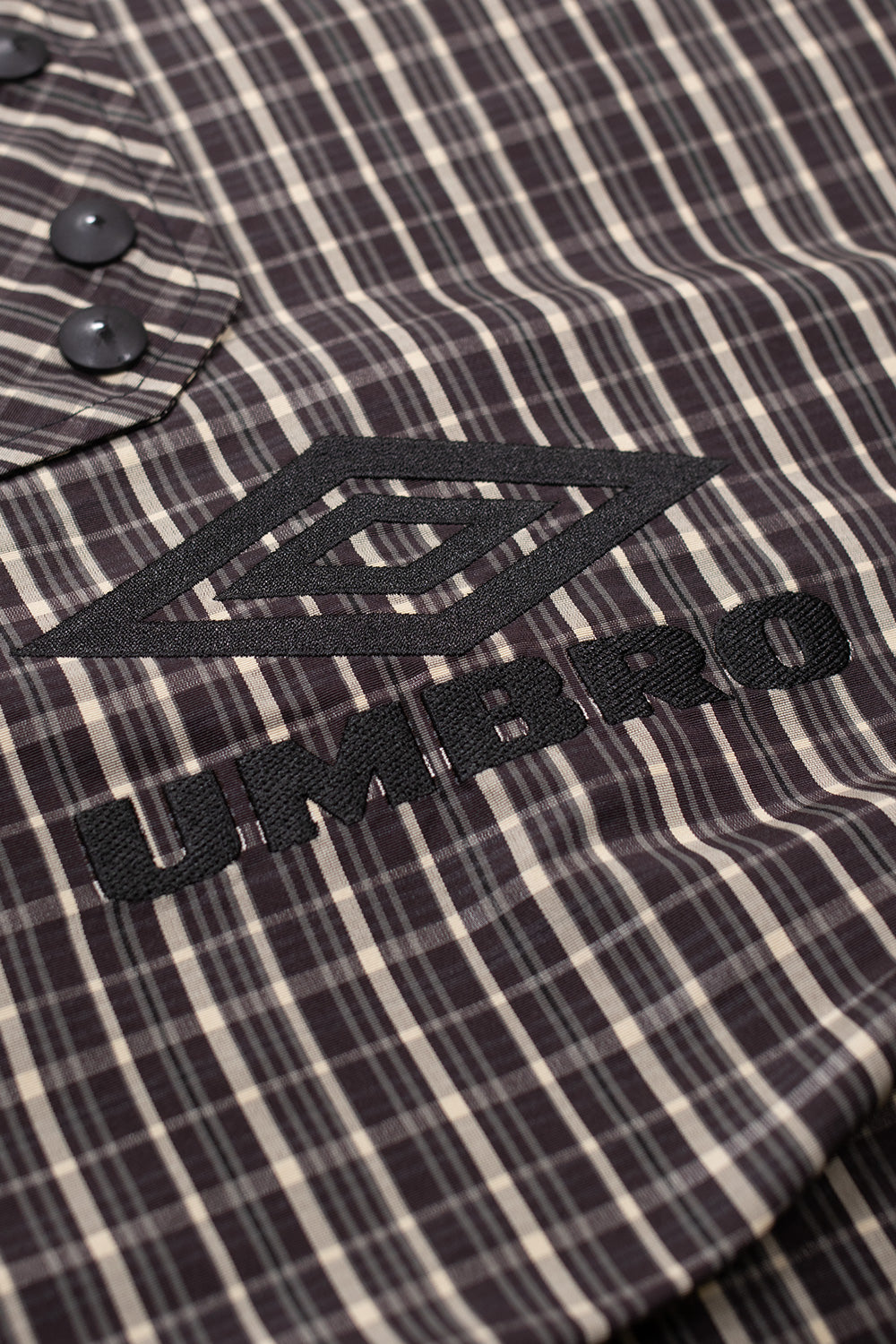 Close-up of embroidered Umbro double diamond logo on beige dark brown check smock anorak, premium streetwear outerwear detail