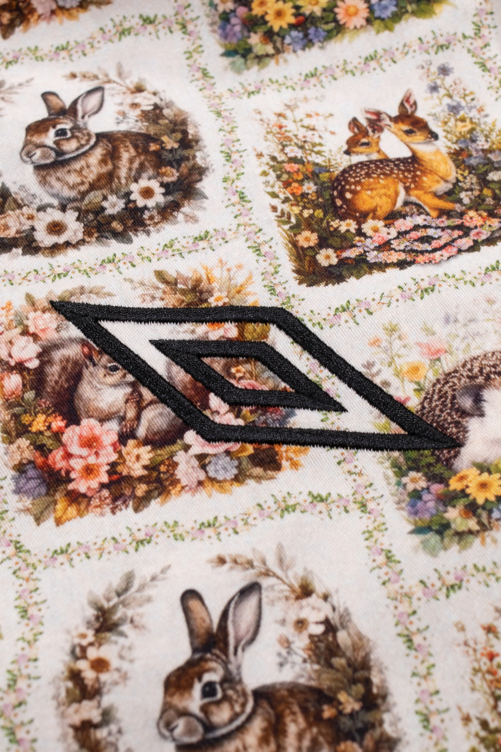 Macro studio close-up of the black satin-embroidered Umbro double diamond logo stitched directly over the textured ivory jacquard fabric, framed by illustrated floral wreaths containing baby fawns, squirrels, and rabbits