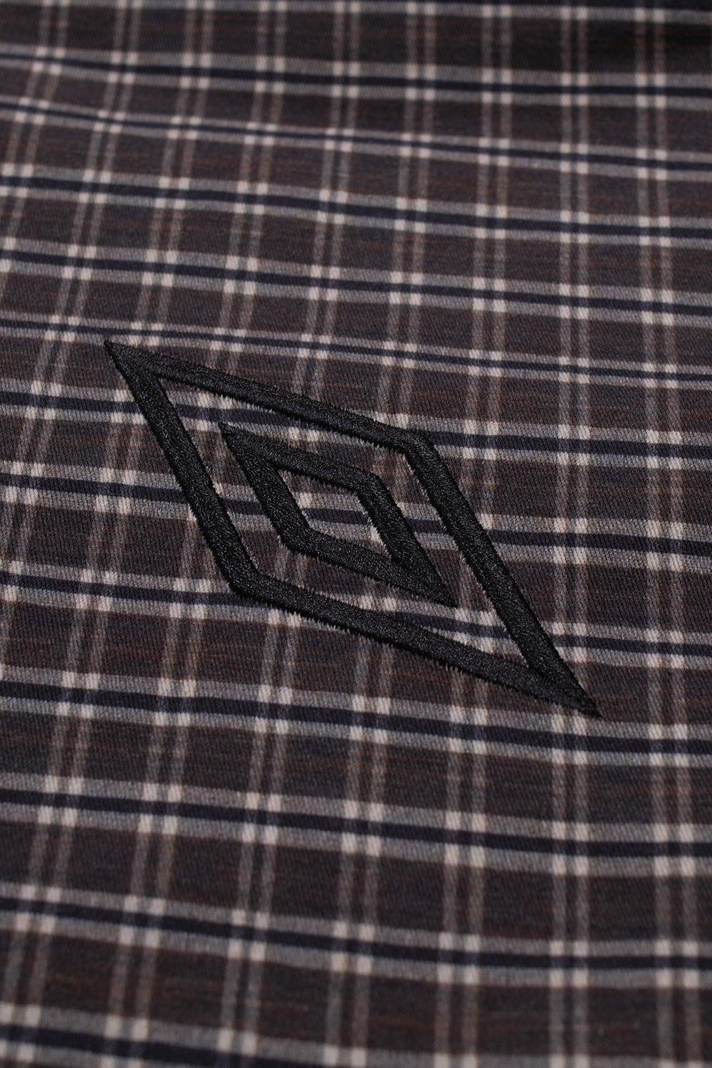 Macro studio close-up of the chest embroidery on the Umbro Double Plaid Jacket, highlighting the clean black satin-stitched Umbro double diamond logo precisely sewn over the textured dark brown tartan cotton fabric