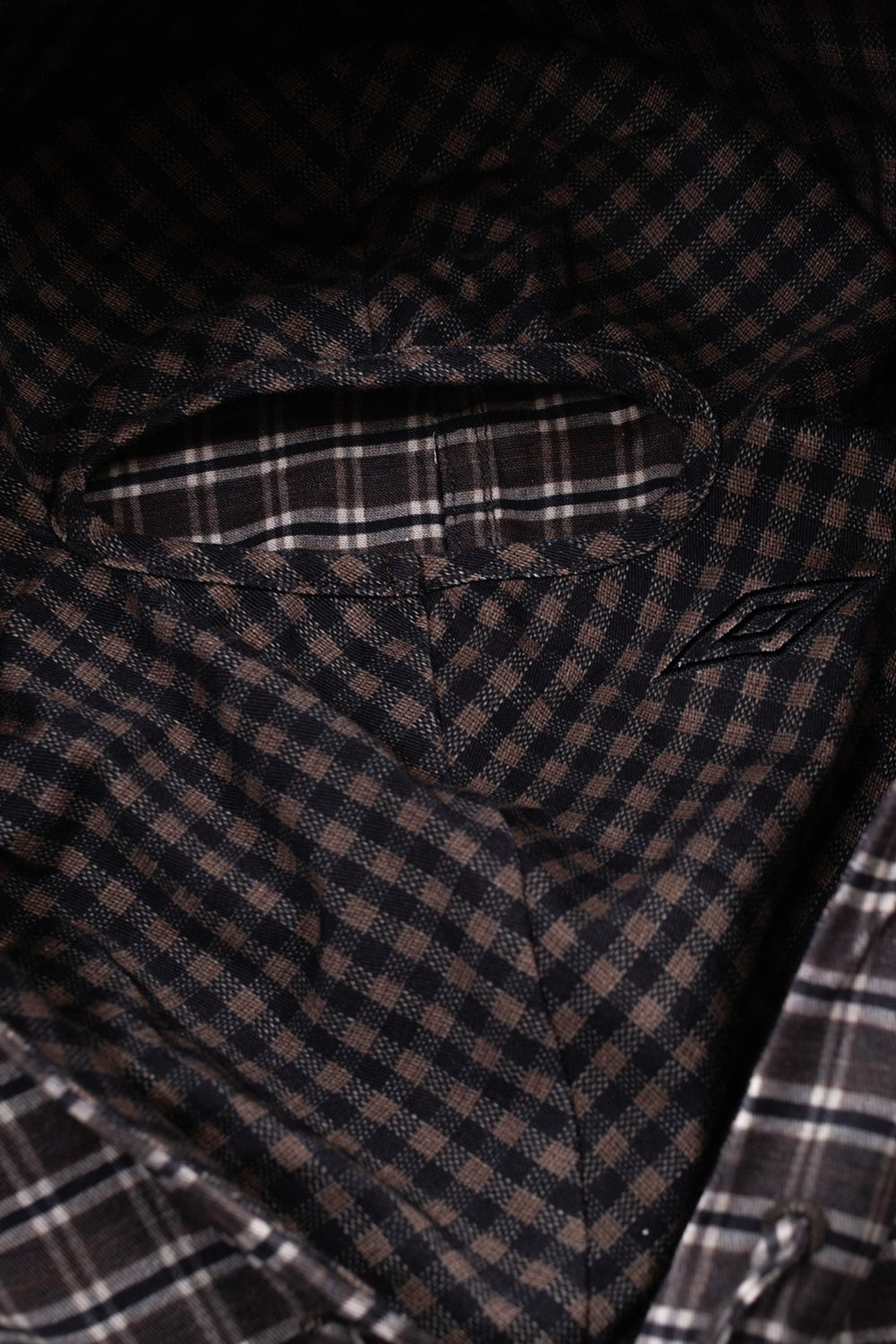 Interior view of the hood on the Umbro Double Plaid Jacket, highlighting the built-in gingham micro-check balaclava face mask panel with a bound horizontal eye opening and a subtle embroidered Umbro diamond accent on the cheek
