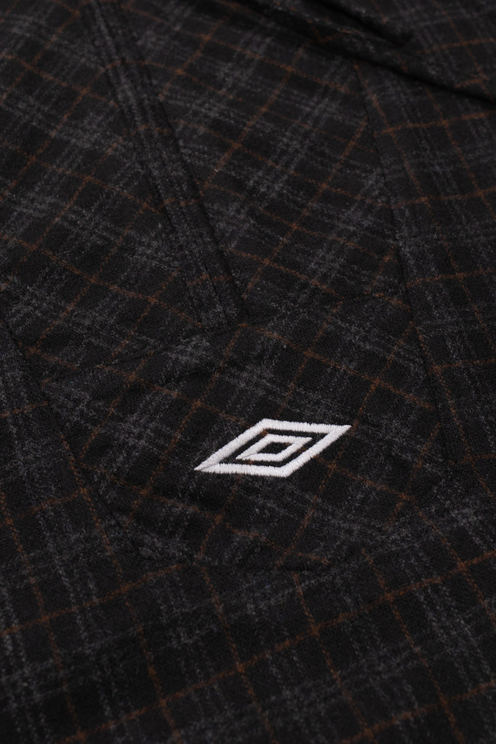 Close-up shot of the front chest detail on the Umbro Flannel Fur Anorak, highlighting the angled flannel placket extension with a white satin-embroidered Umbro double diamond logo over the fuzzy textured dark brown plaid wool blend