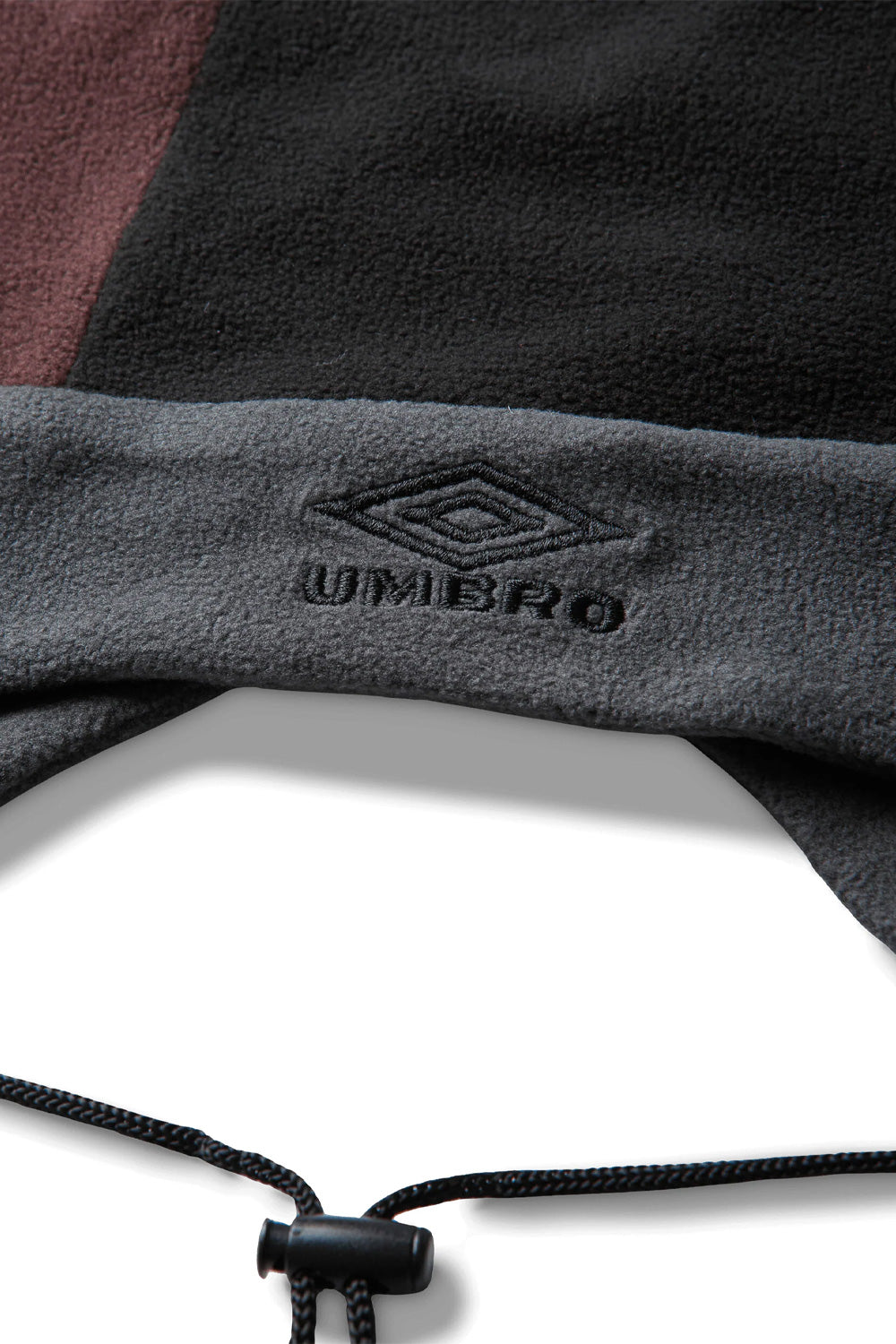 Macro close-up shot of the front band on the Umbro Fleece Jester Hat, detailing the dark charcoal grey embroidery of the iconic Umbro double diamond logo and serif brand lettering on the soft fuzzy charcoal fleece band, accented below by the textured black cord and plastic toggle lock