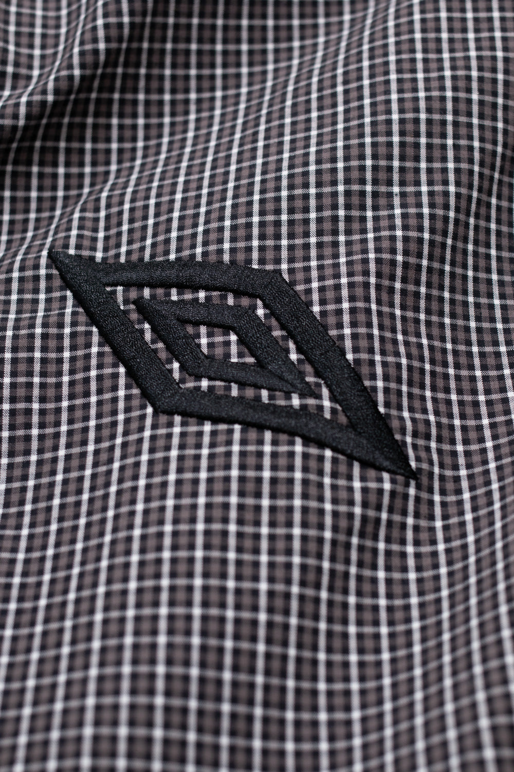 Close-up of embroidered Umbro logo on the Gingham Masked Shirt Coffee Brown / White, emphasizing iconic British sports branding and elevated streetwear craftsmanship.
