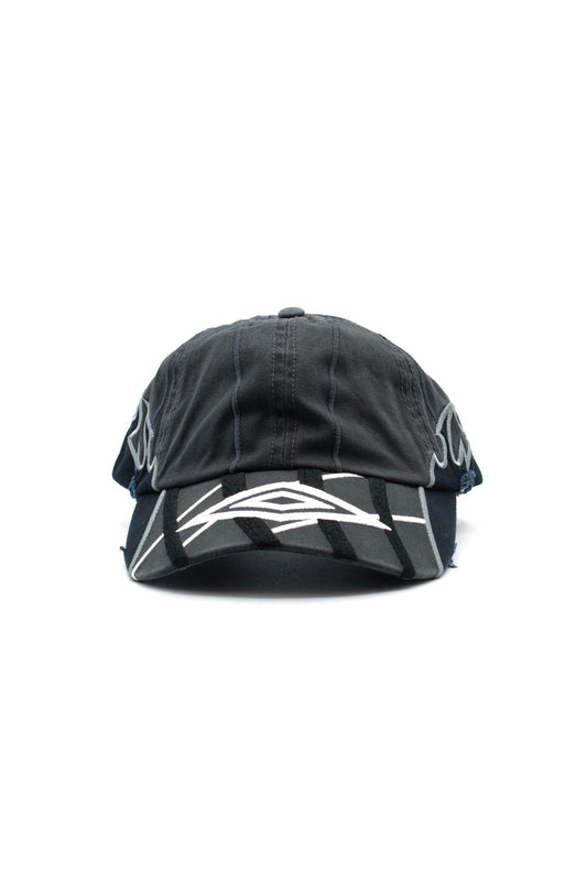 Front studio portrait of the Umbro Goalkeeper Cap in washed dark grey cotton, showcasing an unstructured 6-panel crown with vertical accent stitching, a pre-curved visor embellished with bold white and black layered tribal wave embroidery, distressed frayed details, and silver-grey embroidery wrapping around the temple panels