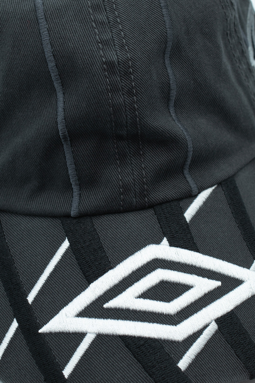 Macro close-up shot of the visor and front panel seam on the Umbro Goalkeeper Cap, detailing the thick white satin-stitched abstract diamond graphics, raised black geometric embroidery, and precise vertical topstitching across the washed dark grey cotton twill