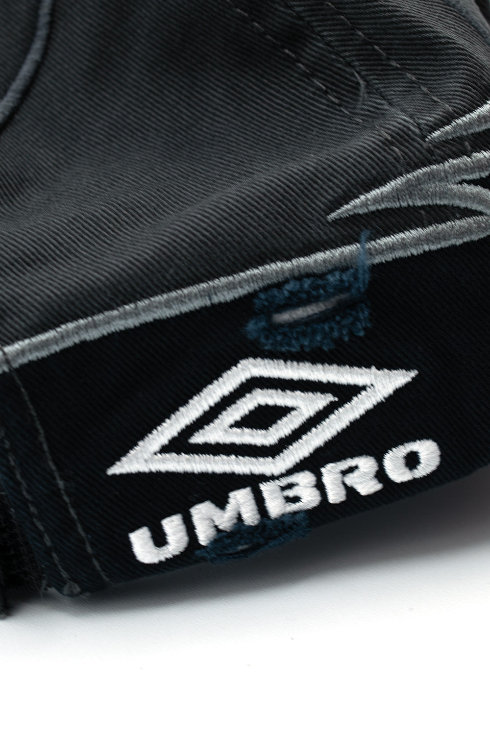 Detailed macro view of the lower rear side panel on the Umbro Goalkeeper Cap, focusing on the dense white satin embroidery of the Umbro double diamond logo and block lettering, accented by subtle distressing and frayed denim-like edge wear