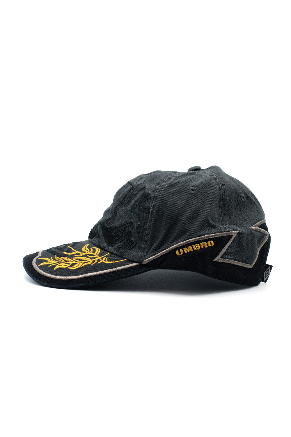 Left profile studio shot of the Umbro Laurel Cap, highlighting the washed dark green cotton twill body, golden yellow embroidered Umbro block lettering along the lower side, curved visor with golden laurel leaf details, and prominent taupe zigzag outline stitching wrapping to the rear