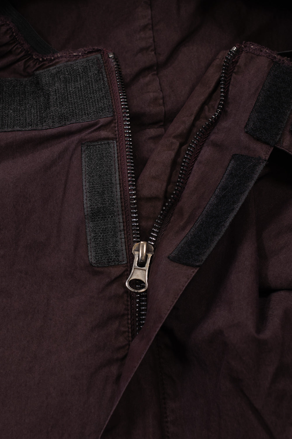 Macro close-up shot of the front closure on the Umbro Midfield Jacket, highlighting the heavy-duty metal zipper track, large metallic pull tab, and wide black hook-and-loop velcro strips securing the protective storm placket on the plum-colored cotton twill fabric