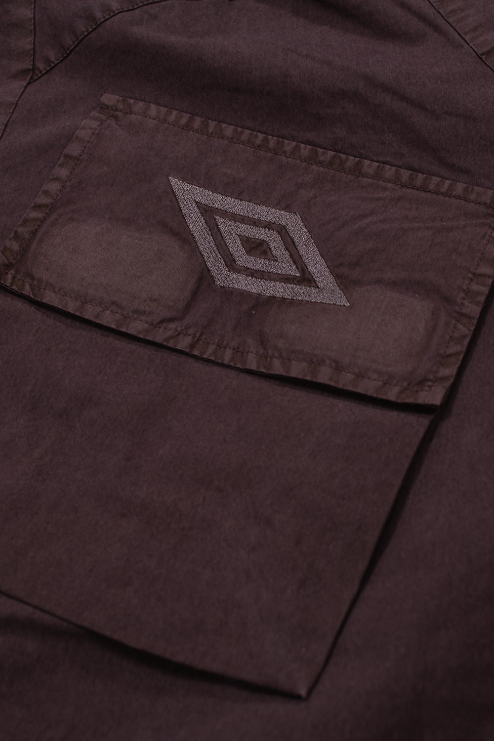Detailed studio close-up of the right angled cargo chest pocket on the Umbro Midfield Jacket, highlighting the reinforced flap with tonal plum satin embroidery of the Umbro double diamond logo over the subtle mineral-washed fabric weave