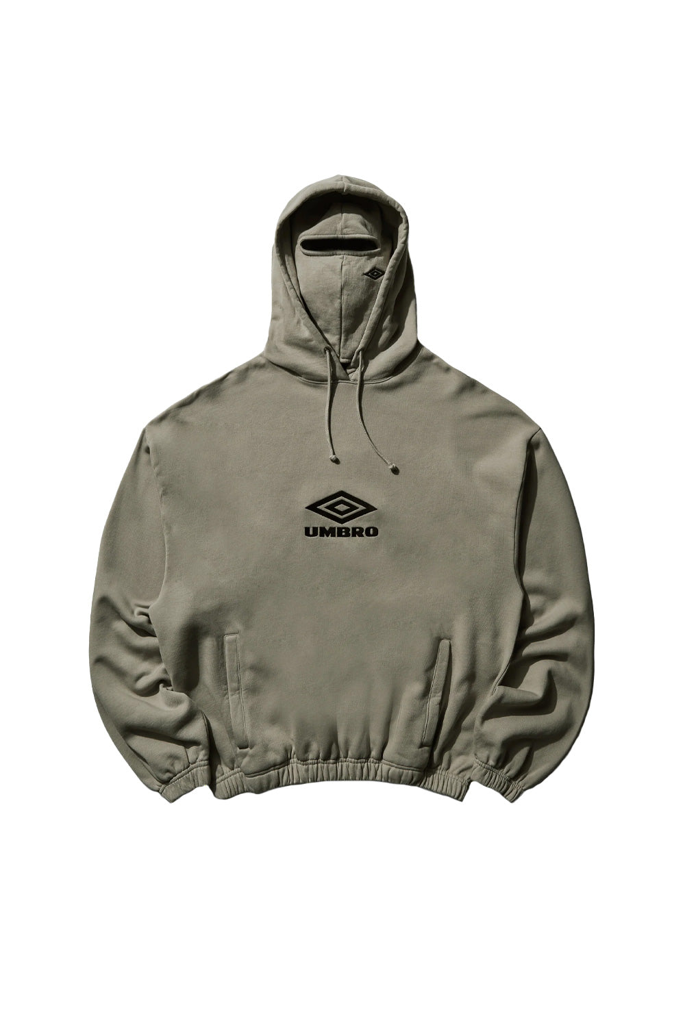 Front flat lay view of the Umbro OG Logo Mask Hoodie in dark greige taupe, showcasing a boxy oversized heavyweight fleece silhouette with elasticated gathered hem and cuffs, vertical side welt pockets, drawstring hood, an integrated matching fabric balaclava mask with a horizontal eye slit, and a black satin-embroidered classic Umbro double diamond wordmark logo across the center chest