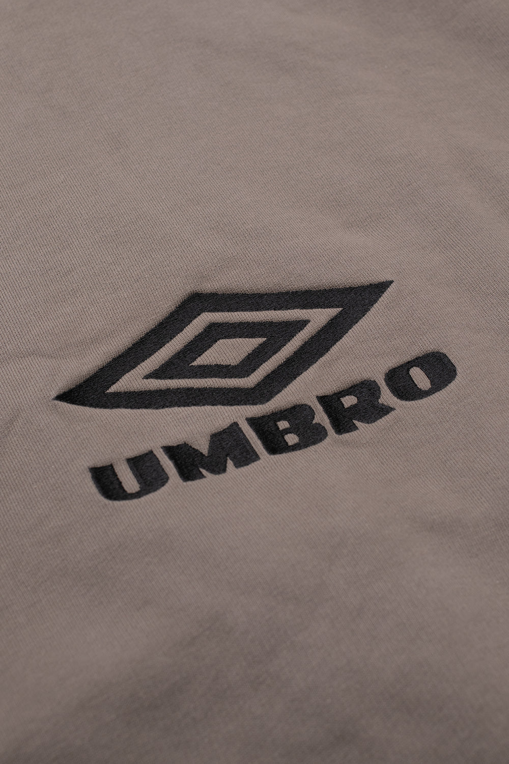 Macro close-up shot of the front chest embroidery on the dark greige Umbro OG Logo Mask Hoodie, detailing the dense black satin stitching of the iconic double diamond outline emblem and bold "UMBRO" brand lettering over the smooth cotton fleece fabric