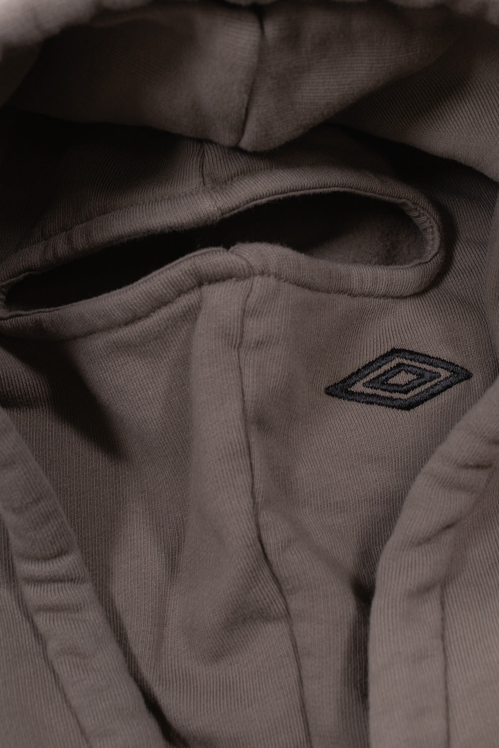 Interior view of the hood on the Umbro OG Logo Mask Hoodie, highlighting the built-in dark greige balaclava face mask panel with a bound horizontal eye opening and a small tonal embroidered Umbro diamond logo positioned on the cheek